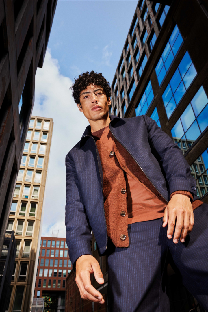 Man in a navy pinstripe jacket and rust knit between city buildings